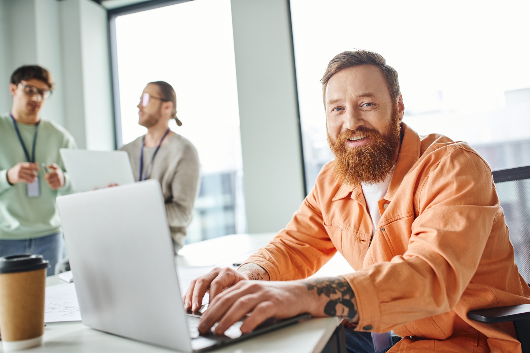 cheerful bearded and tattooed architect working on laptop near takeaway drink in paper cup and looking at camera while colleagues talking on blurred background in coworking design studio Cheerful Bearded And Tattooed Architect Working On MATHSH5